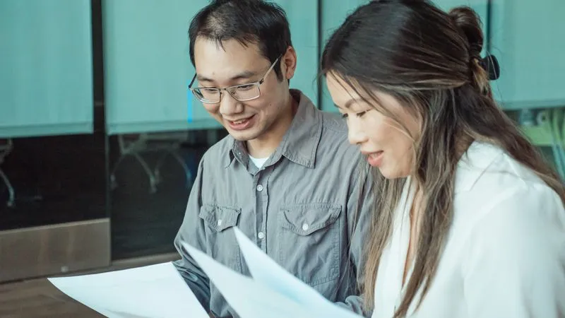 Two smiling colleagues look over printed documents together in an office.