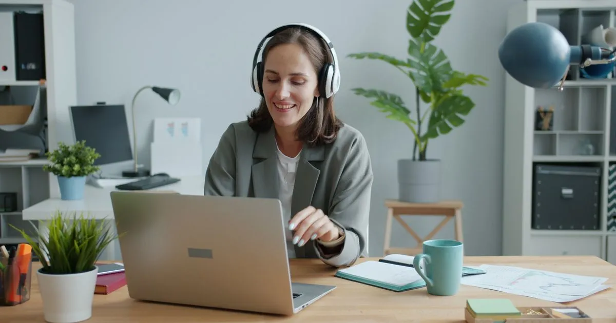 Smiling woman wearing headphones types on a laptop at a wooden desk in an office.