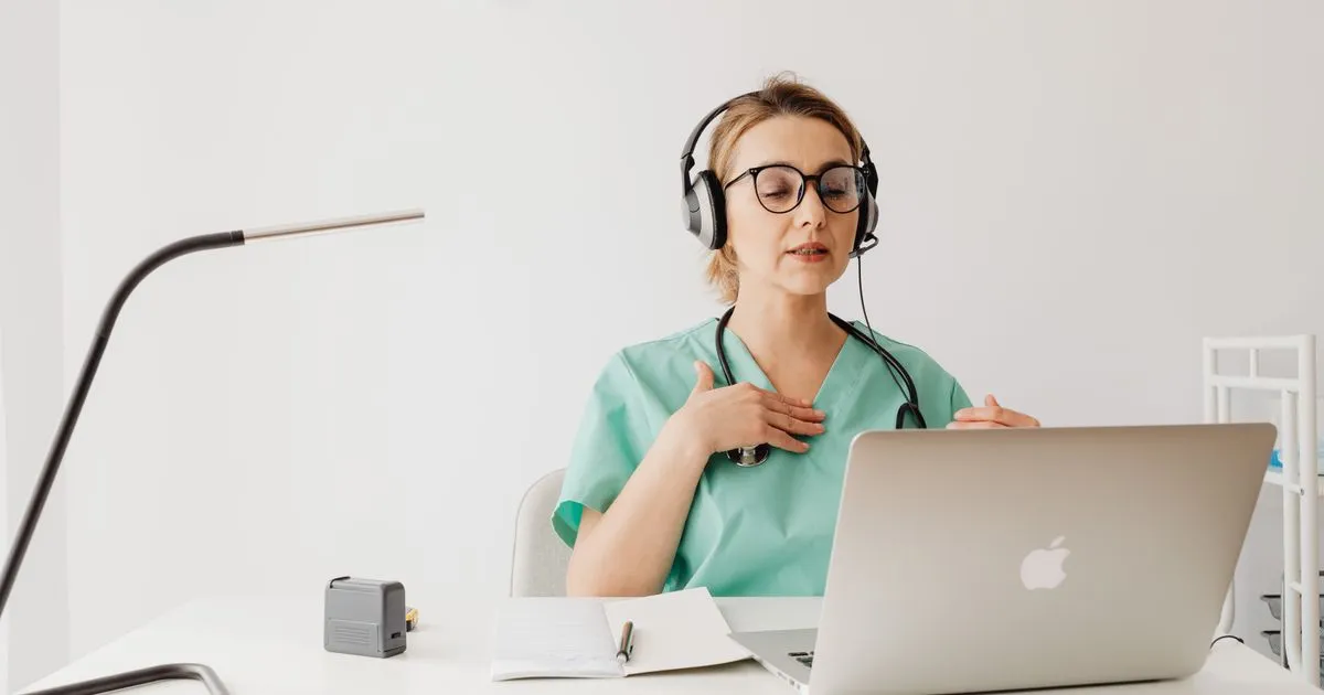 Healthcare worker in green scrubs and a headset speaking during a laptop video call.