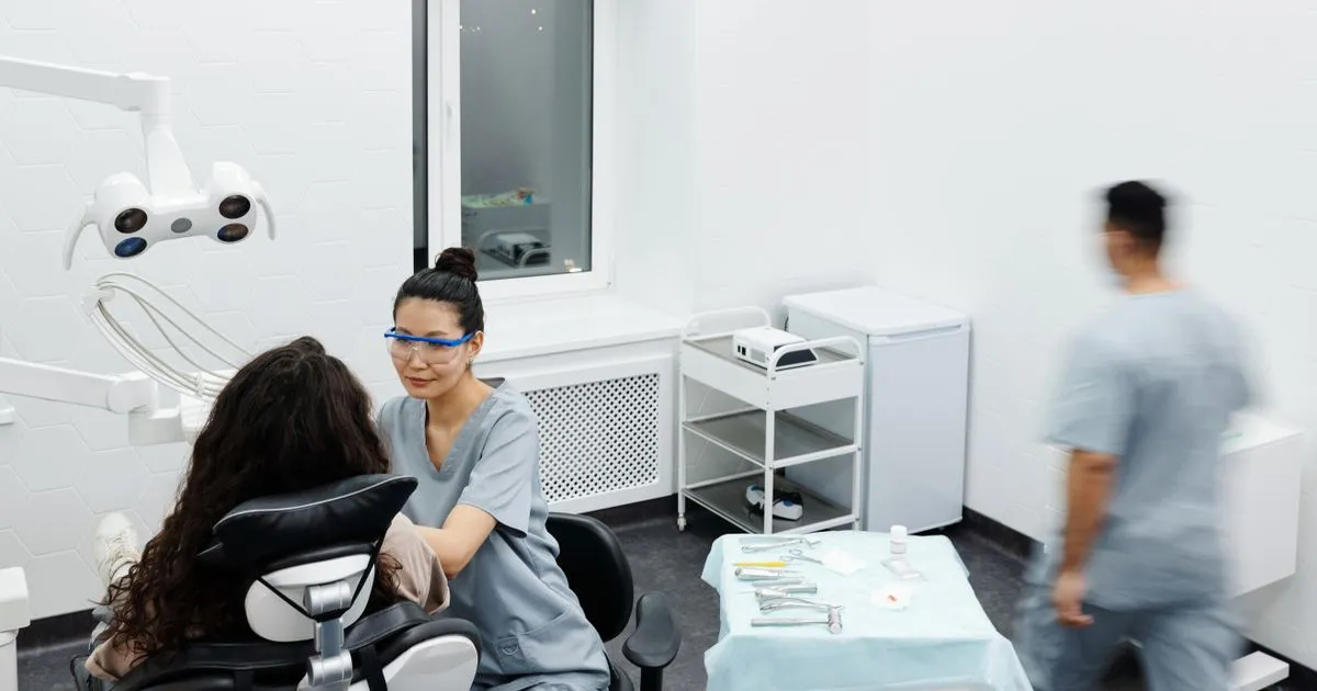 A dental worker in safety glasses talks with a patient seated in a dental chair.
