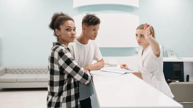 Receptionist in a white uniform points the way for two visitors at a clinic front desk.