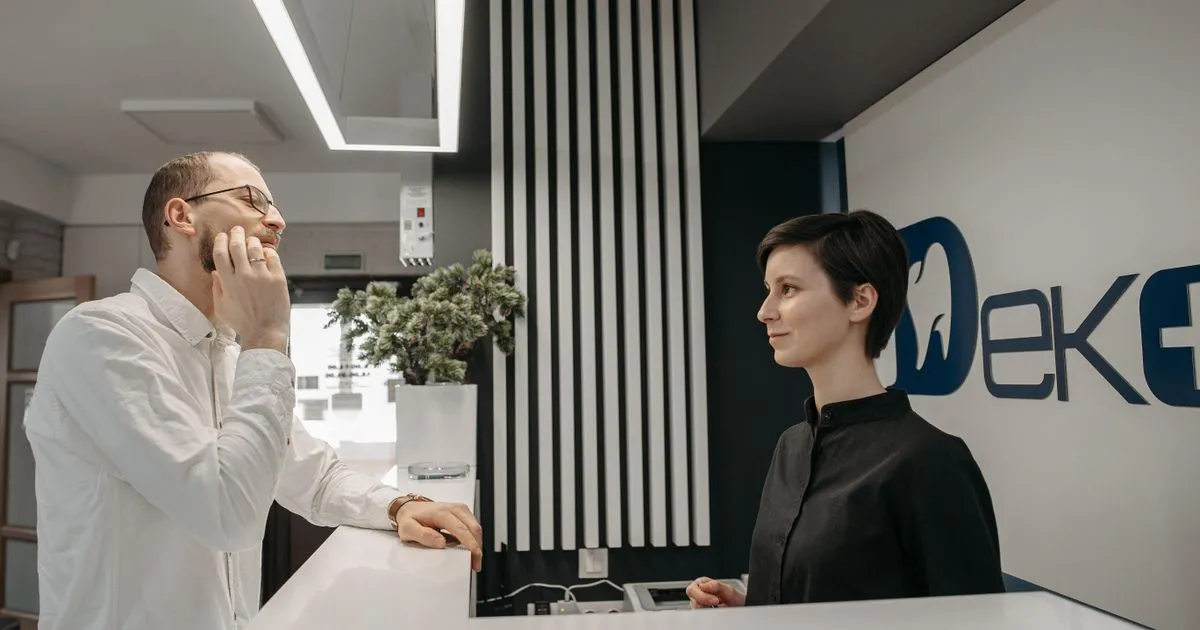 A man touching his cheek talks with a receptionist across a white clinic front desk.
