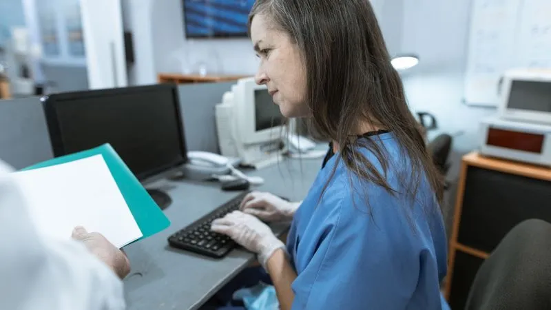 Healthcare worker in blue scrubs and gloves typing at a computer while a colleague holds papers.