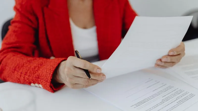 A person in a red jacket reads printed documents at a desk, holding a pen.