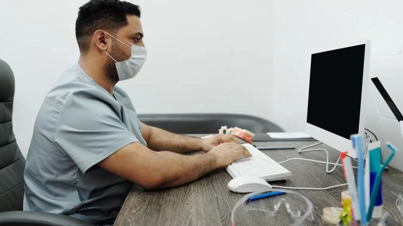 Man in scrubs and a face mask typing on a computer at a dental office desk.