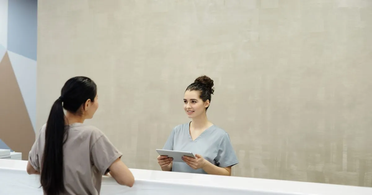 A smiling staff member in scrubs holds a tablet while talking with a woman at a reception desk.