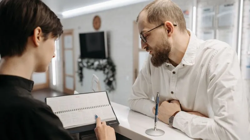 A receptionist points to a desk calendar while talking with a man at a reception counter.