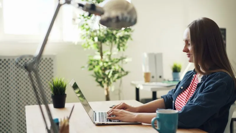 Woman smiling while typing on a laptop at a bright desk with plants.