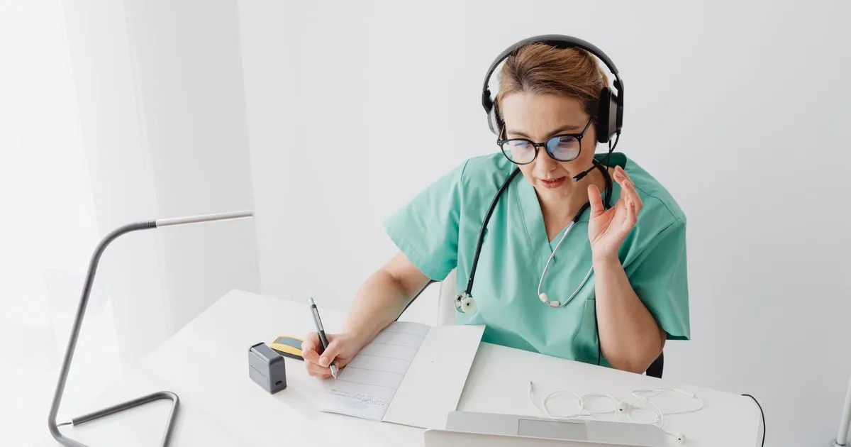 Healthcare worker in scrubs and a headset writes notes at a desk with a laptop.