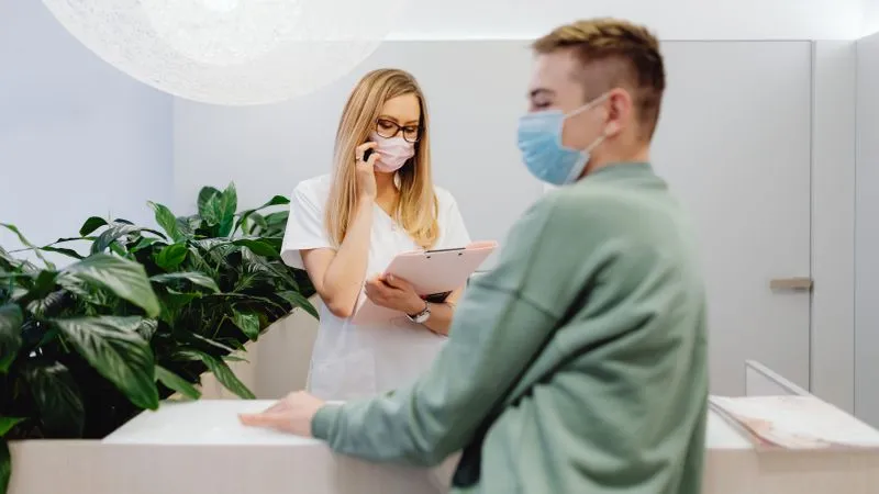 Masked clinic receptionist talks on the phone holding a clipboard while a masked patient waits at the desk.
