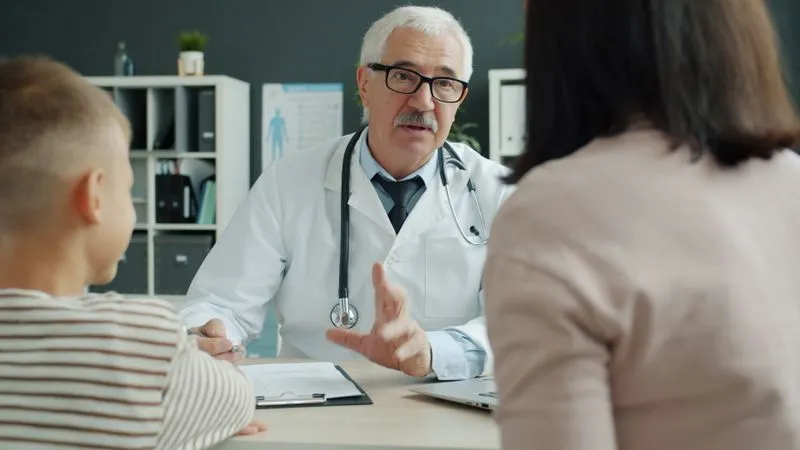 A doctor in a white coat talks with a woman and a child across a desk.