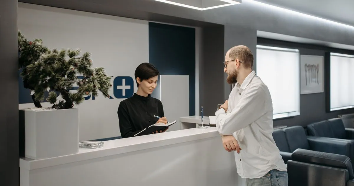 A receptionist takes notes at a clinic front desk while a man talks with her.