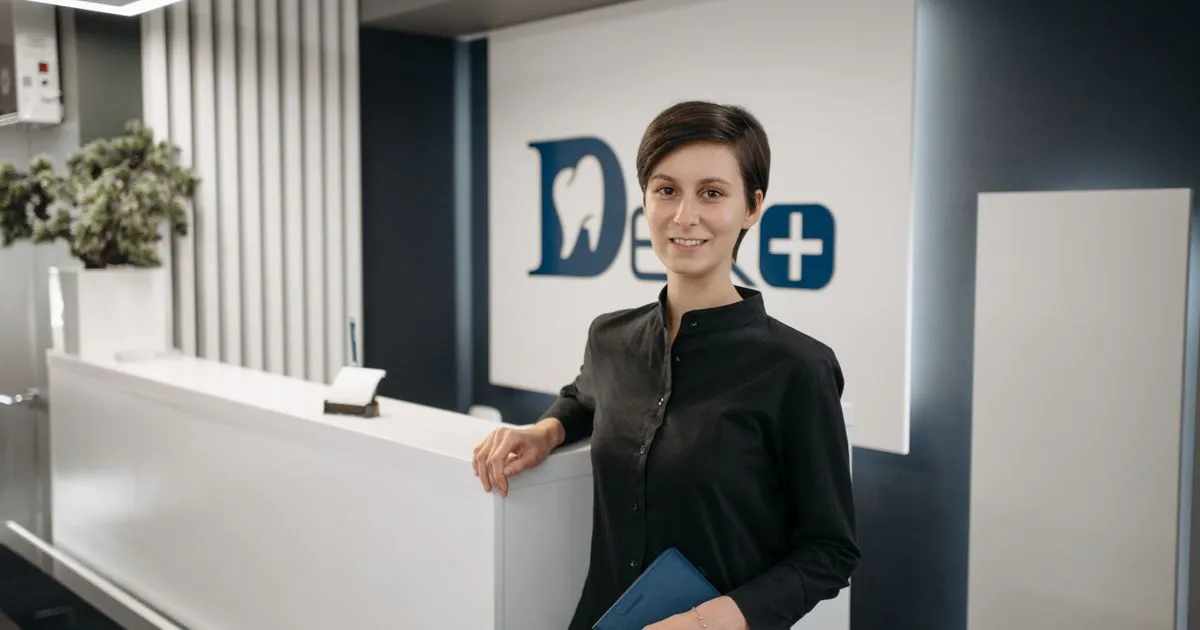 Smiling woman in a black shirt standing beside a white reception desk in a dental clinic.
